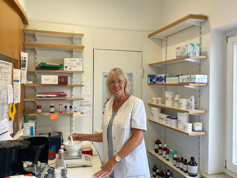 Eine Apotheken-Mitarbeiterin sieht in die Kamera und stellt eine Rezeptur her, mit der Hand an einem Mörser.